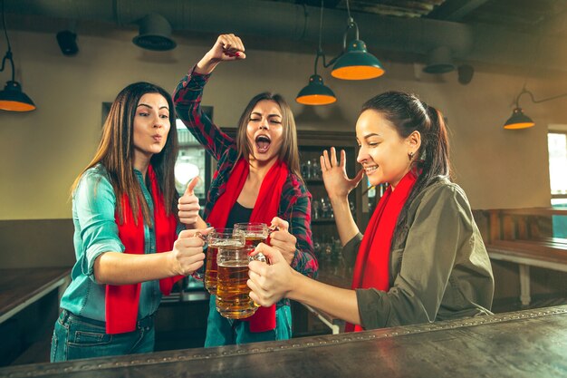 Sport, people, leisure, friendship, entertainment concept - happy female football fans or good young friends drinking beer, celebrating victory at bar or pub. Human positive emotions concept