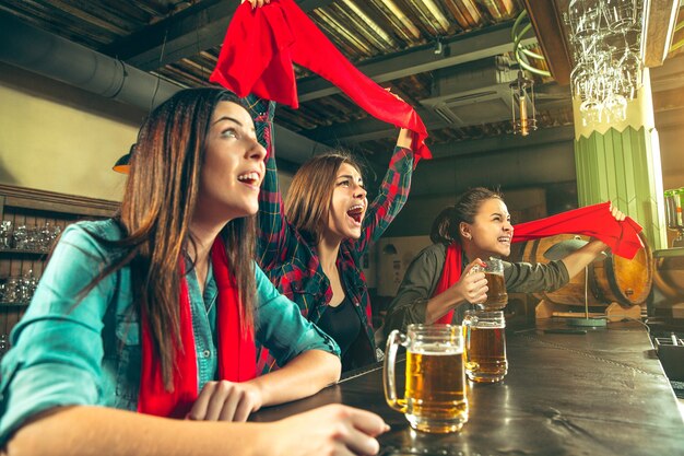 Free photo sport, people, leisure, friendship, entertainment concept - happy female football fans or good young friends drinking beer, celebrating victory at bar or pub. human positive emotions concept
