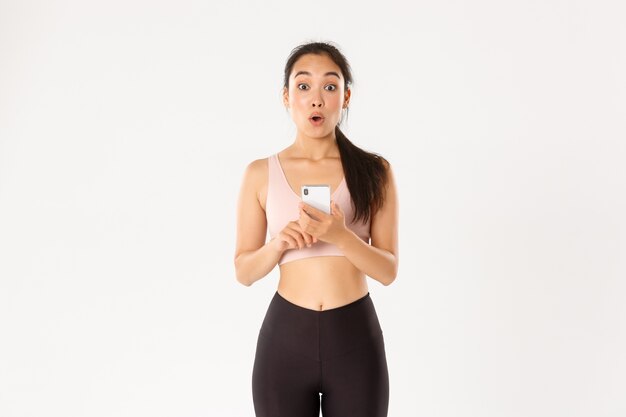 스포츠, 기술 및 활동적인 라이프 스타일 개념. 놀랍고 놀란 아시아 여성 운동 선수 인 운동가는 휴대 전화에서 피트니스 앱을 확인하고 운동 추적기를 사용하여 응용 프로그램을 실행하면서 와우라고 말합니다.