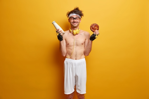 Sport and weight loss concept. Positive sportsman has temptation to eat bun with milk, breaks diet, feels hungry after exercising in gym, wears sports clothes, isolated over yellow wall