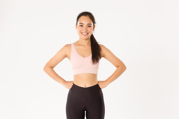 Sport, wellbeing and active lifestyle concept. Cheerful slim and strong asian fitness girl workout in gym, standing in activewear with hands on waist, smiling pleased after good training in gym.