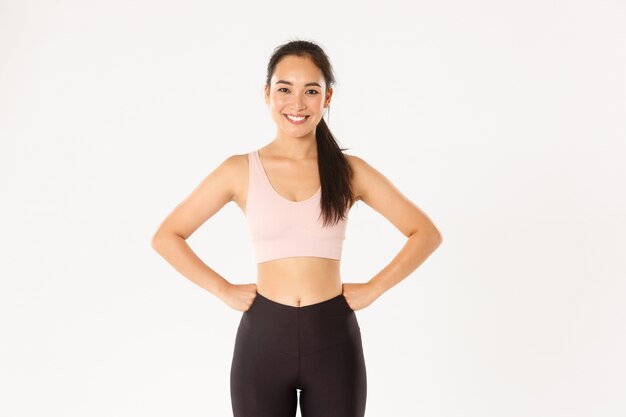 Sport, wellbeing and active lifestyle concept. Cheerful slim and strong asian fitness girl workout in gym, standing in activewear with hands on waist, smiling pleased after good training in gym.