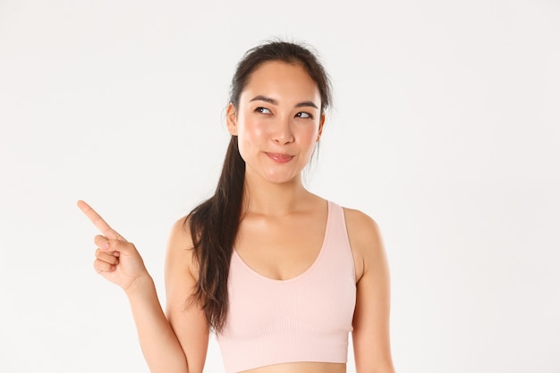 Sport, wellbeing and active lifestyle concept. Cunning and thoughtful smiling asian sportswoman, fitness girl making her choice, looking pleased with intrigued smile, pointing upper left corner.