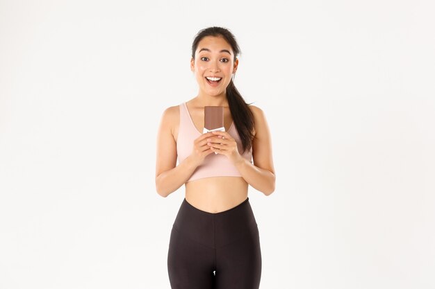 Sport, wellbeing and active lifestyle concept. Happy smiling asian female athlete holding chocolate protein bad and looking excited, eating healthy sweets for prolonged workout.