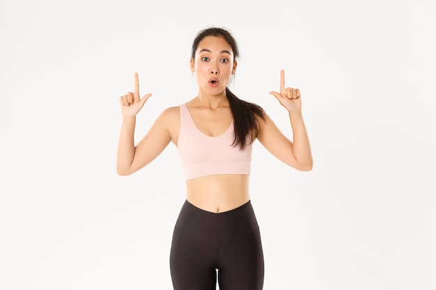 Sport, wellbeing and active lifestyle concept. Impressed and curious asian female athlete saying wow, looking amazed and pointing fingers up, want to know discount and special offers details.