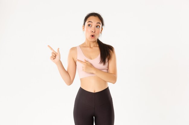 Sport, wellbeing and active lifestyle concept. Impressed and excited sportswoman, asian athlete in activwear open mouth wondered and pointing fingers upper left corner at your logo banner.