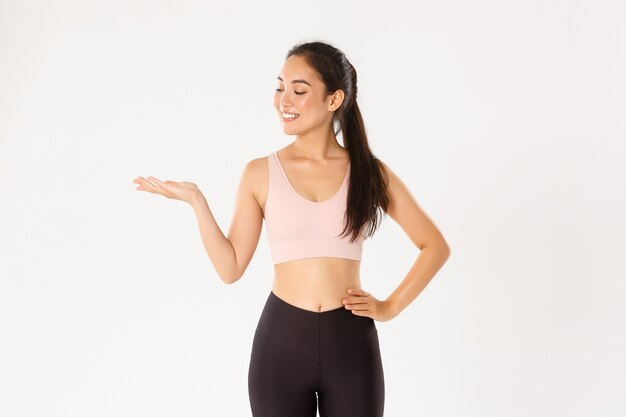 Sport, wellbeing and active lifestyle concept. Portrait of cheerful attractive asian fitness coach, female athlete introduce or demonstrate product for workout, holding something on hand.