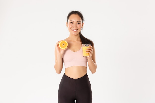 Sport, wellbeing and active lifestyle concept. Portrait of smiling healthy and slim asian girl advice eating healthy food for breakfast, gain energy for workout, hold fresh juice and orange