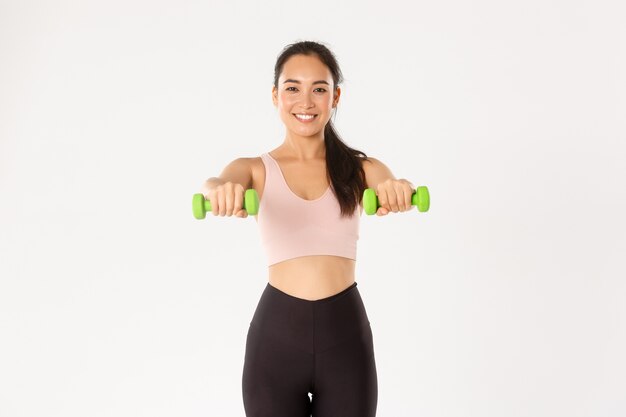 スポーツ ジム 健康的な体のコンセプト 笑顔の女性アスリートの全長 ダンベルでフィットネス運動をしているかわいい女の子 ジム 白い背景でアクティブウェアトレーニングのスポーツウーマン プレミアム写真