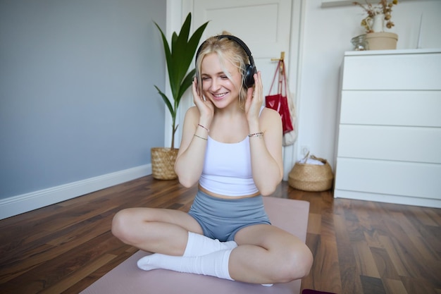 Free photo sport and wellbeing concept smiling woman in wireless headphones sitting on yoga mat listens to