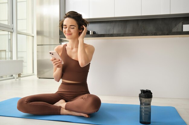 スポーツと女性のライフスタイルの若い女性は、明るいところでスマートフォンを使ってヨガマットに座って自宅でトレーニングを行う