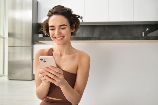 Sport and women lifestyle young woman does workout at home sits on yoga mat with smartphone in brigh