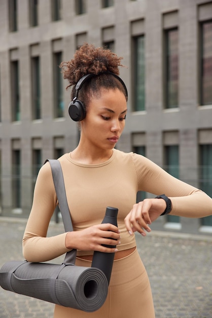 Sport and workout concept Active woman dressed in beige tracksuit checks fitness results carries fitness mat bottle of water exercises outdoors poses against modern city building listens music