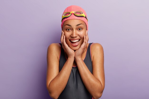 Sportive cheerful dark skinned girl wears black bodysuit, swimcap and swimglasses