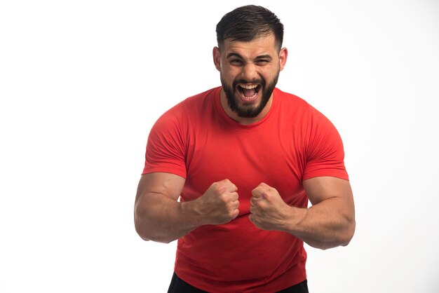 Sportive man in red shirt demonstrating his arm muscles and screaming. 