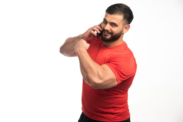 Sportive man in red shirt talking to the phone and demonstrating his arm muscles . 