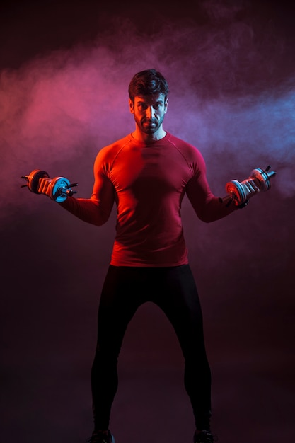 Sportive man standing with dumbbells in dark