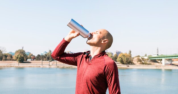 Sportive young man drinking water from bottle standing near the lake