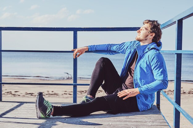 Sportive young man is chilling near sea while sitting on balcony at bright sunny day.