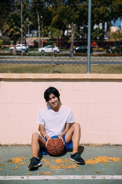 Sportive young man sitting on asphalt with basketball outdoors