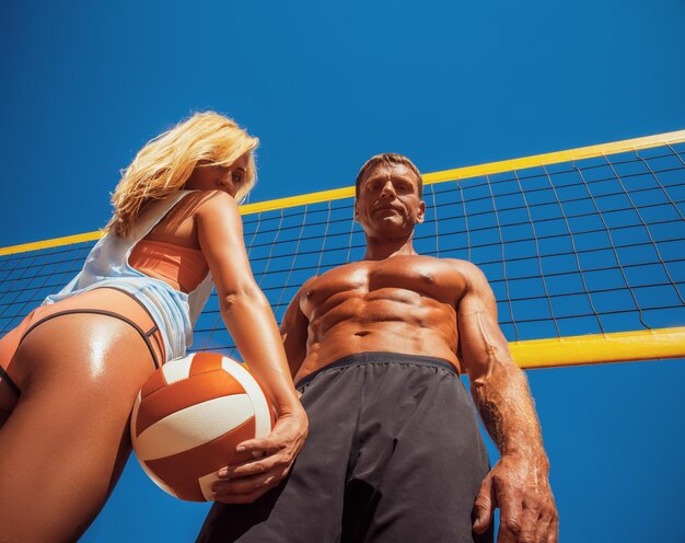 Sports couple. Handsome muscular tanned male and sexy tanned blonde girl, standing on a volleyball ground.