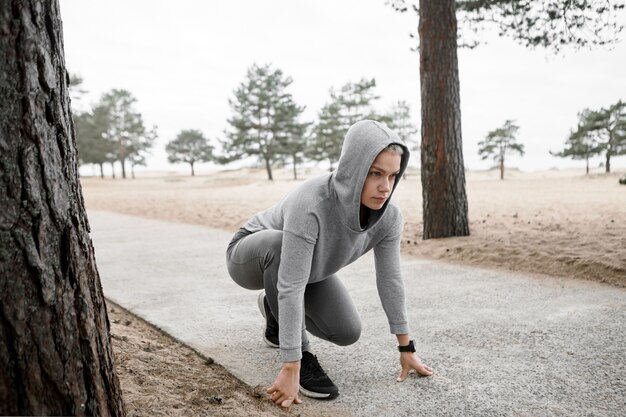 Sports, fitness, wellness, health, energy and competition concept. Outdoor image of concentrated young female athlete in hoodie and sneakers sitting in steady position on paved trail, ready to run