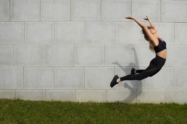 Спорт, здоровье, активность, фитнес, благополучие и летняя концепция. Замороженный снимок красивой молодой кавказской спортсменки в стильной черной одежде, высоко прыгающей во время разминки на открытом воздухе