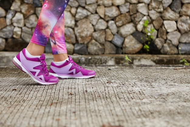 Спорт и здоровый образ жизни. Закройте ноги женщины в стильных фиолетовых кроссовках и леггинсах с космическим принтом на тротуаре. Спортсменка, стоящая на бетоне, делает физические упражнения в городском парке