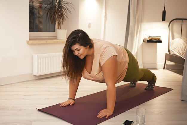 Sports, healthy lifestyle, fitness and obesity concept. Energetic self determined young plus size female in sportswear doing plank exercise on mat. Curvy brunette woman exercising at home, planking