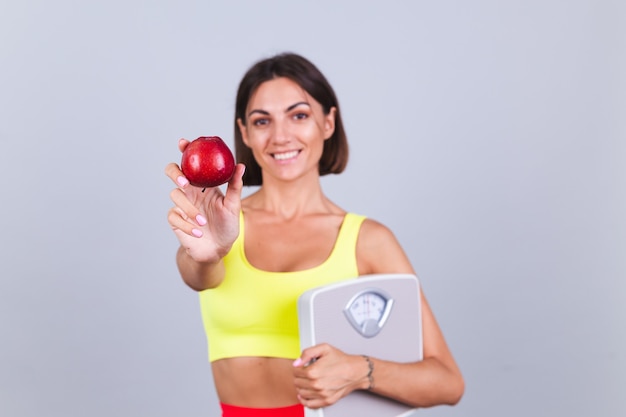 Sports woman stands on gray wall, satisfied with the results of fitness training and diet, holding scale, wears top and leggings, holds apple