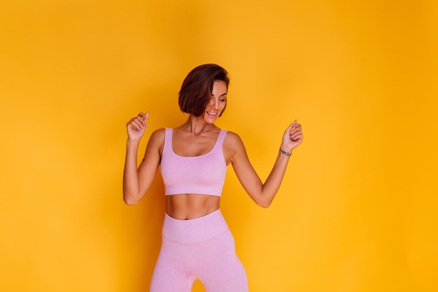 Free photo sports woman stands on yellow wall, demonstrating her abs, satisfied with the results of fitness training and diet, has a happy facial expression, wears a sports top and tight leggings