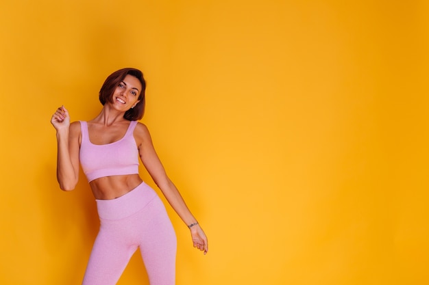 Sports woman stands on yellow wall, demonstrating her abs, satisfied with the results of fitness training and diet, has a happy facial expression, wears a sports top and tight leggings