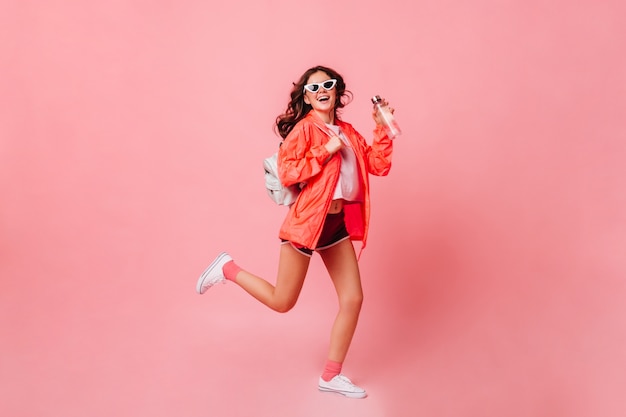 Sports woman in windbreaker, shorts and sneakers runs on pink wall