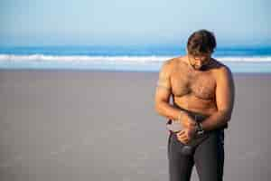 Free photo sportsman putting on wetsuit for surfing on ocean beach and taking off watch