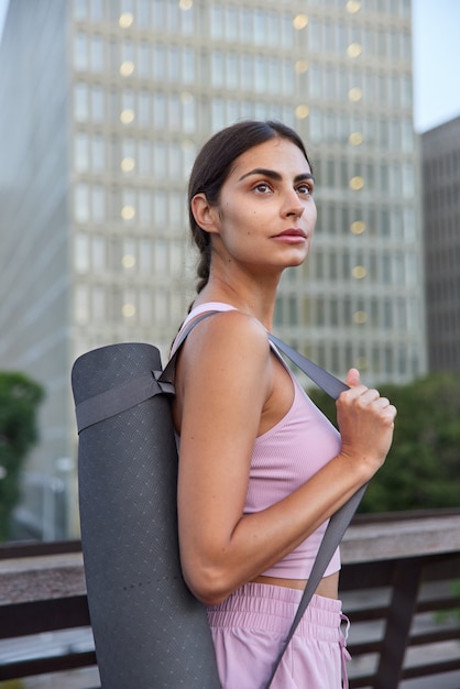  sportswoman in activewear carries rolled mat on shoulder going to have exercises in open air concentrated away poses on city scrapers