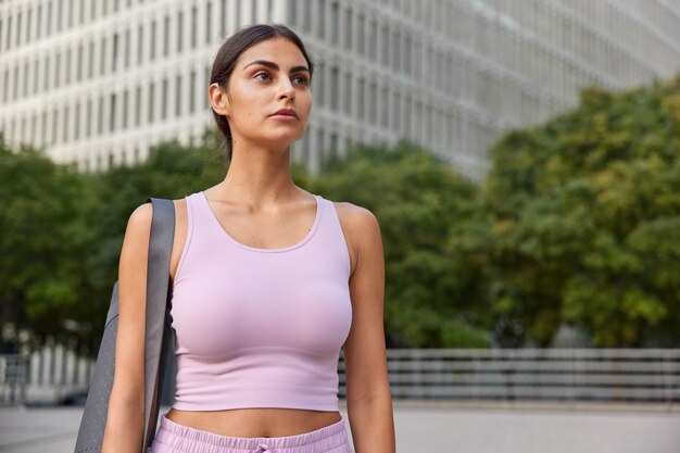  sportswoman being in good physical shape carries mat on shoulder leads active lifestyle poses in urban settings strolls outdoors returns after workout