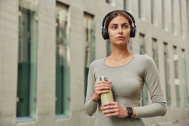 Free photo sportswoman dressed in acitivewear holds bottle of water recreats after cardio training uses wireless headphones looks away poses near modern city building