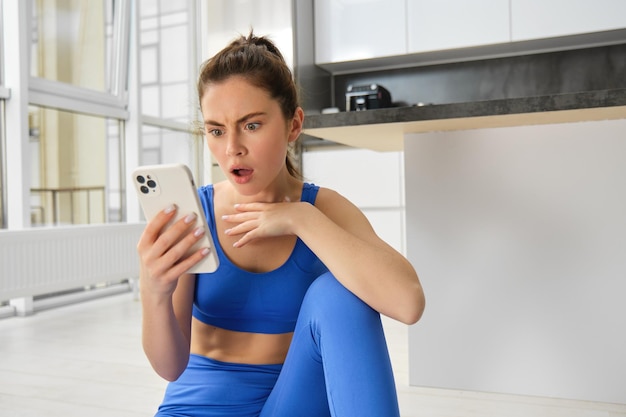 Free photo sportswoman looking at horrible news on smartphone screen sitting in middle of living room at home