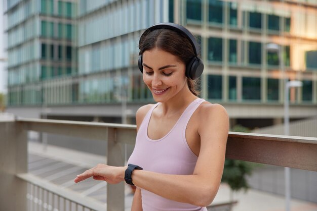 Sporty active woman in casual t shirt looks at smartwatch focused at screen smiles gladfully satisfied after wokout listens music in wireless headphones poses outdoors against urban surroundings