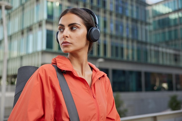 Sporty active woman concentrated into distance with thoughtful expression carries rolled fitness mat gets ready for training poses outdoors against city background. Athletic female exercises regularly