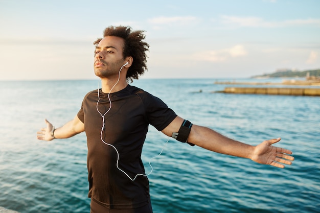 Sporty afro-American man stretching his arms before outdoor workout. Slim and strong male athlete wearing black T-shirt, keeping his arms open, breathing fresh sea air.