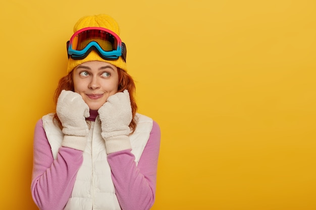 Sporty athletic girl skier looks pensively aside, wears white winter gloves and vest, snowboarding goggles, looks aside, poses against yellow studio wall, blank space