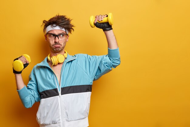 スポーティなアスレチックストロングマンはダンベルを持ち上げ、上腕二頭筋のトレーニングに一生懸命働き、アクティブで健康的なライフスタイルを導き、定期的な運動をし、黄色い壁にポーズをとり、空きスペースを脇に置きます