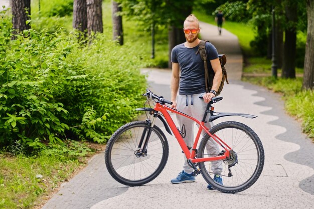 スポーティなひげを生やした男性は、屋外の赤いマウンテンバイクに座っています。