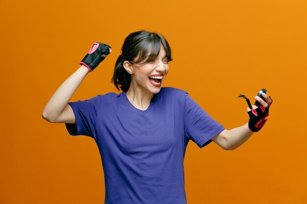 Спортивная красивая женщина в спортивной одежде в перчатках, держащая секундомер, смотрит на него, счастливая и взволнованная, сжимая кулак, стоя на оранжевом фоне