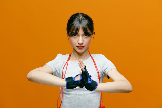 Sporty beautiful woman in sportswear with jumping rope in gloves holding hands together looking at camera with serious face standing over orange background