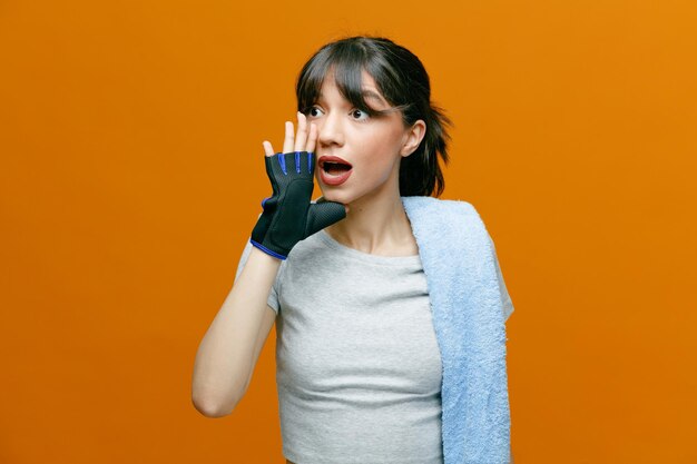 Sporty beautiful woman in sportswear with towel on her shoulder in gloves keeps hand near mouth like calling someone standing over orange background