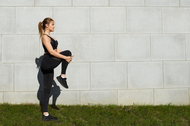 朝の公園でポニーテールとアスレチックボディトレーニングをしているスポーティなブロンドの女の子、有酸素運動の後に脚の筋肉を伸ばし、テキストのコピースペースで灰色のレンガの壁の背景に対してポーズをとる