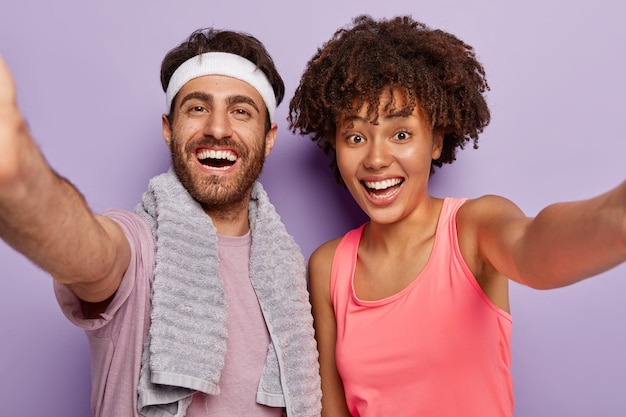 Free photo sporty couple makes selfie after fitness exercise, smile broadly, express good emotions, wear casual clothes, keep hands extended, lead healthy lifestyle, isolated over purple wall. workout, training