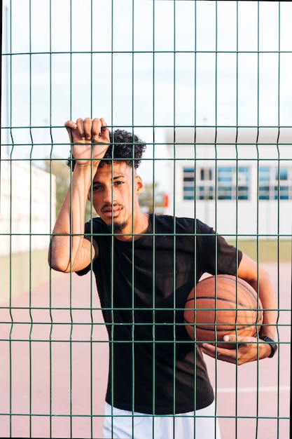 Sporty ethnic man holding basketball behind fence